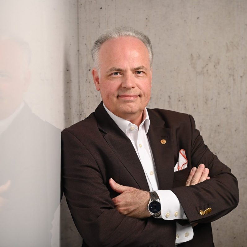 Männlicher Psychotherapeut mit verschränkten Armen, grauem Haar und braunem Sakko vor Betonwand.